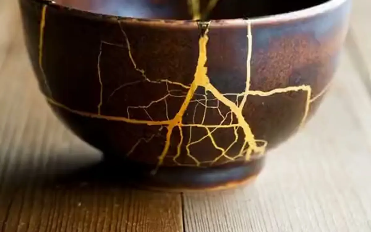 A dark brown tea bowl on a wooden table, its cracks mended with bright gold seams.